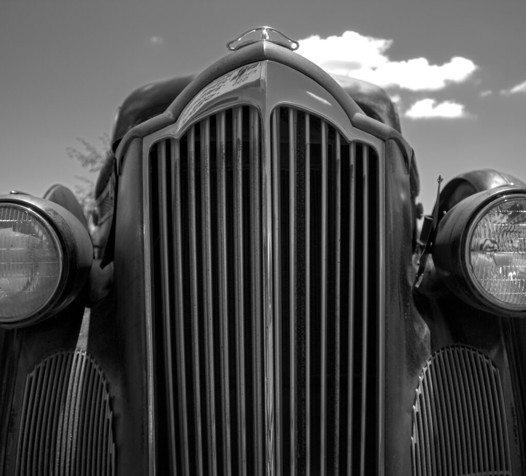 “Black and white photo of a classic car’s front view, showcasing a vertical grille and rounded headlights.”