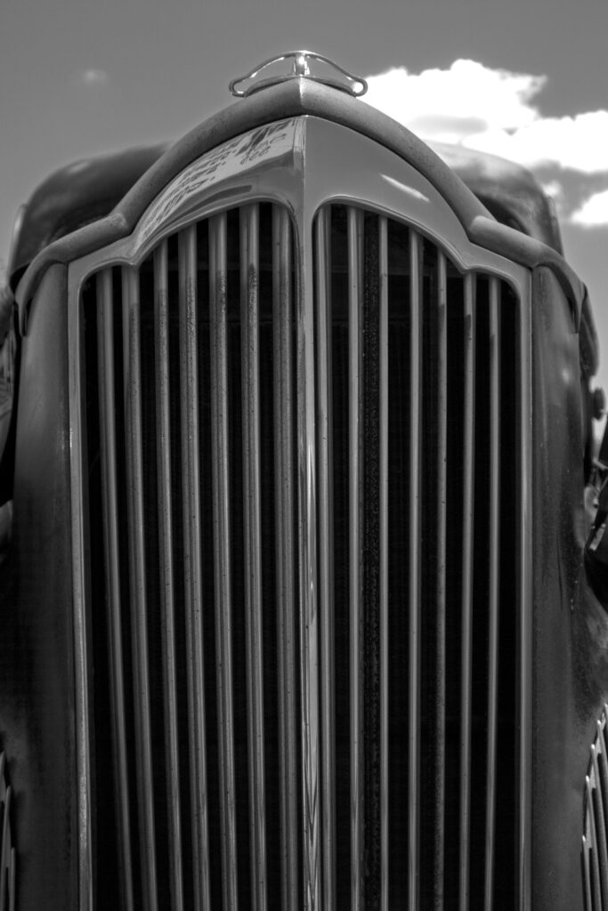 “Black and white photo of a classic car’s front view, showcasing a vertical grille and rounded headlights.”