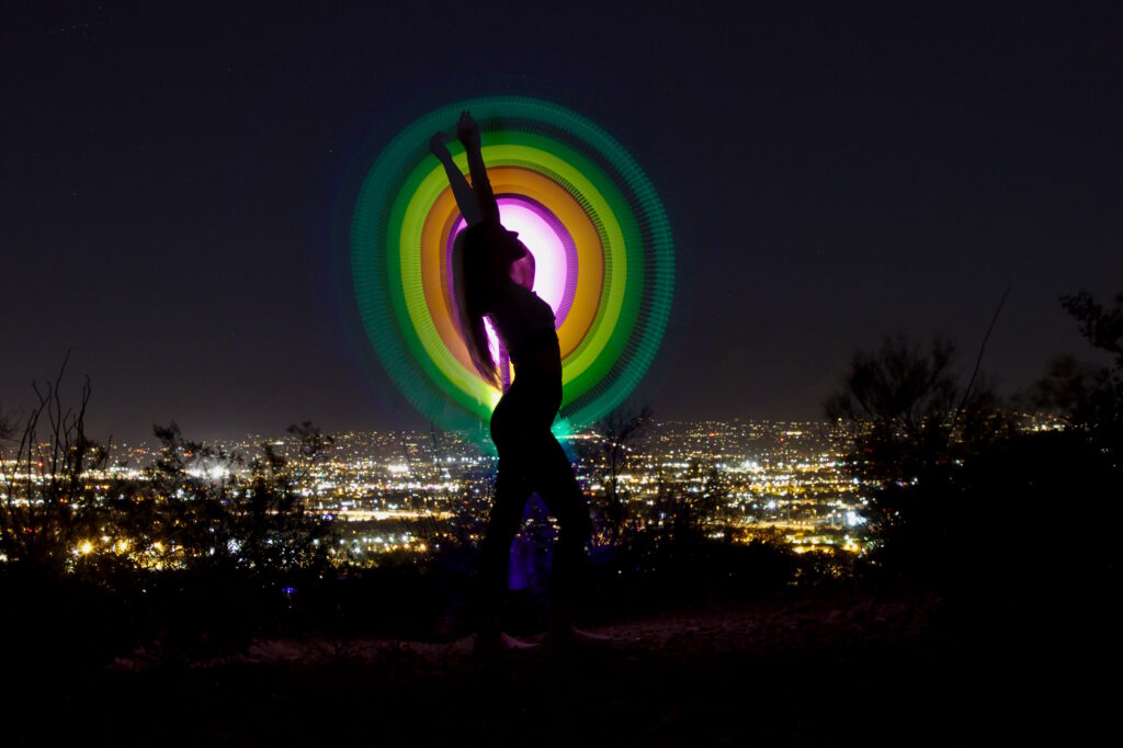 “Silhouette against a vibrant circular light painting under the night sky, with city lights in the distance.”